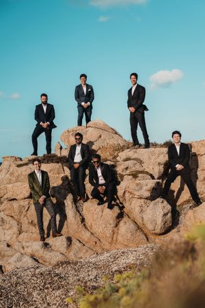 Groomsmen in dark suits pose dramatically on desert rock formation against blue sky in Sardinia.