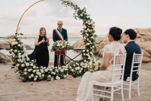 Outdoor wedding ceremony setup with floral arch and white chairs against rocky coastal backdrop, in Sardinia.