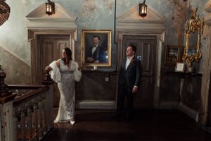 Elegant couple in formal attire pose in grand staircase area of historic building with vintage portraits at Paradise by Kensal Rise.