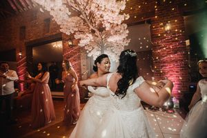 Two people in wedding dresses dance joyfully at a reception with floating confetti and pink uplighting.