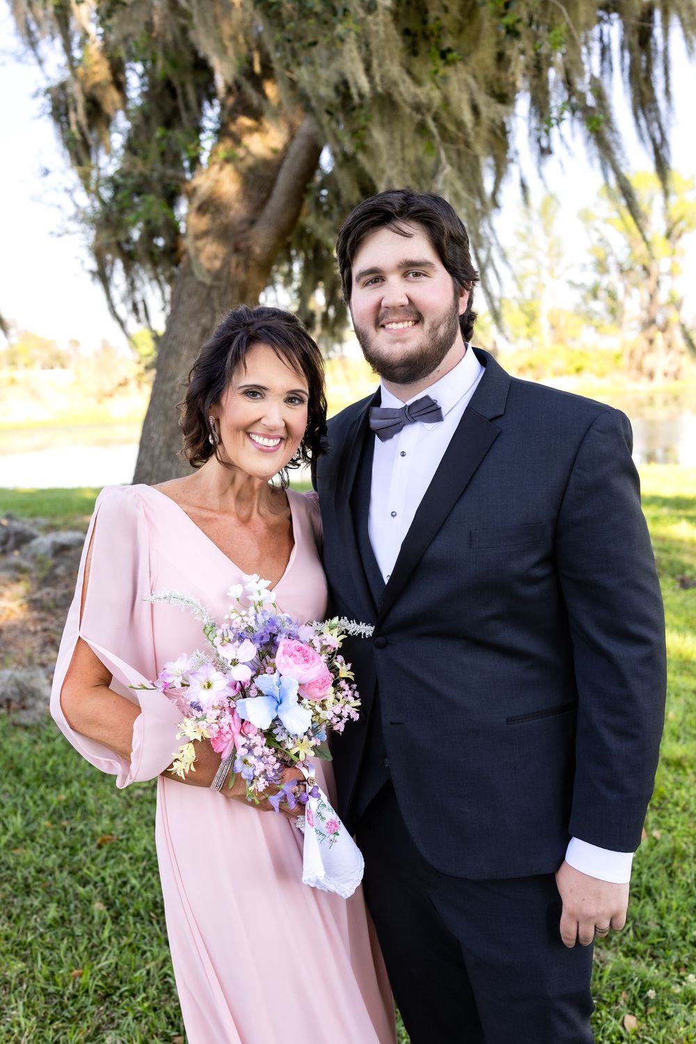 Woman in pink gown holding floral bouquet poses with man in black tuxedo at outdoor wedding celebration.