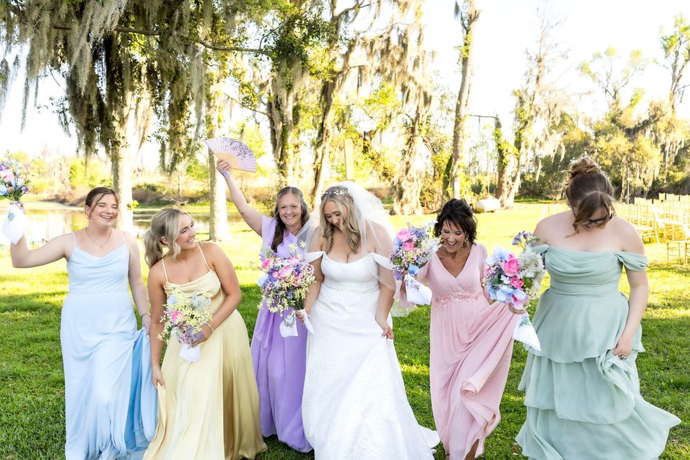 Bride and bridesmaids in colorful mismatched dresses celebrating outdoors under Spanish moss-draped trees.