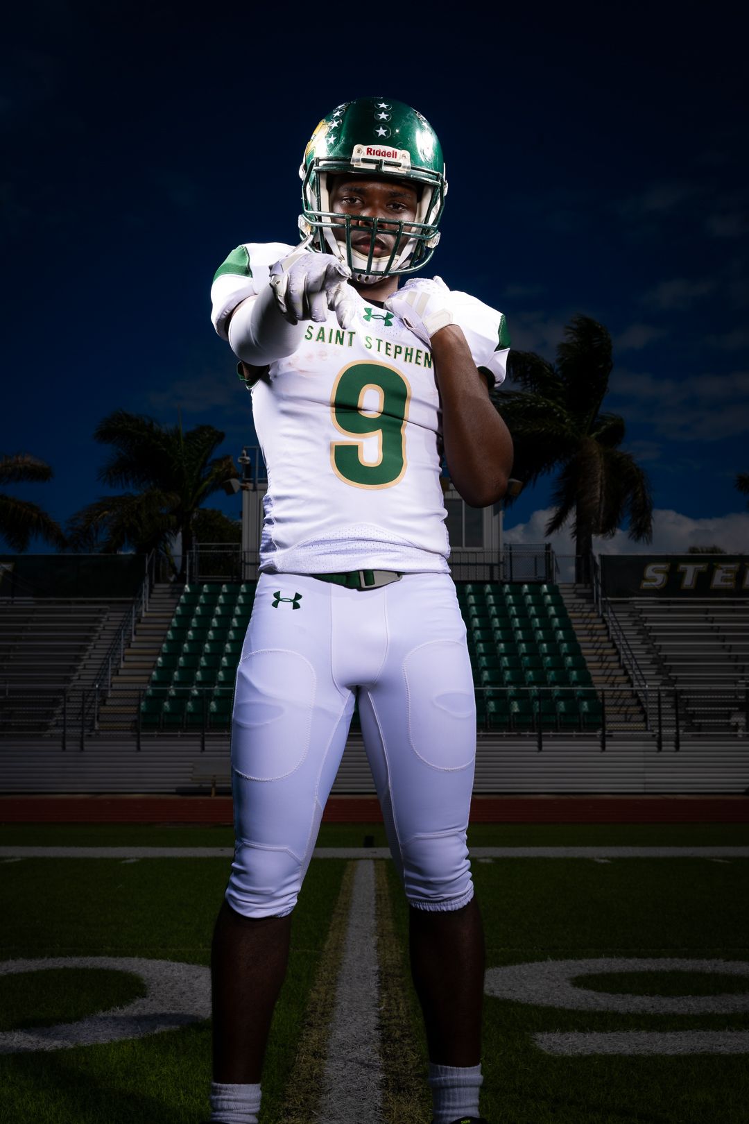 Football player in white and green uniform poses on field at night with stadium lights.