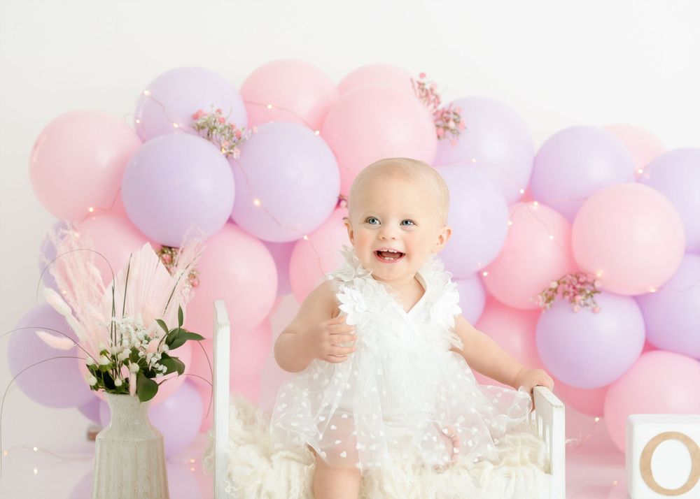 Smiling baby in white dress with pink and purple balloon arch backdrop.