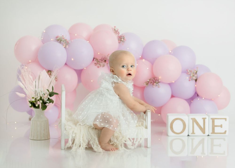 First birthday photoshoot with white lace dress against pastel balloon wall.