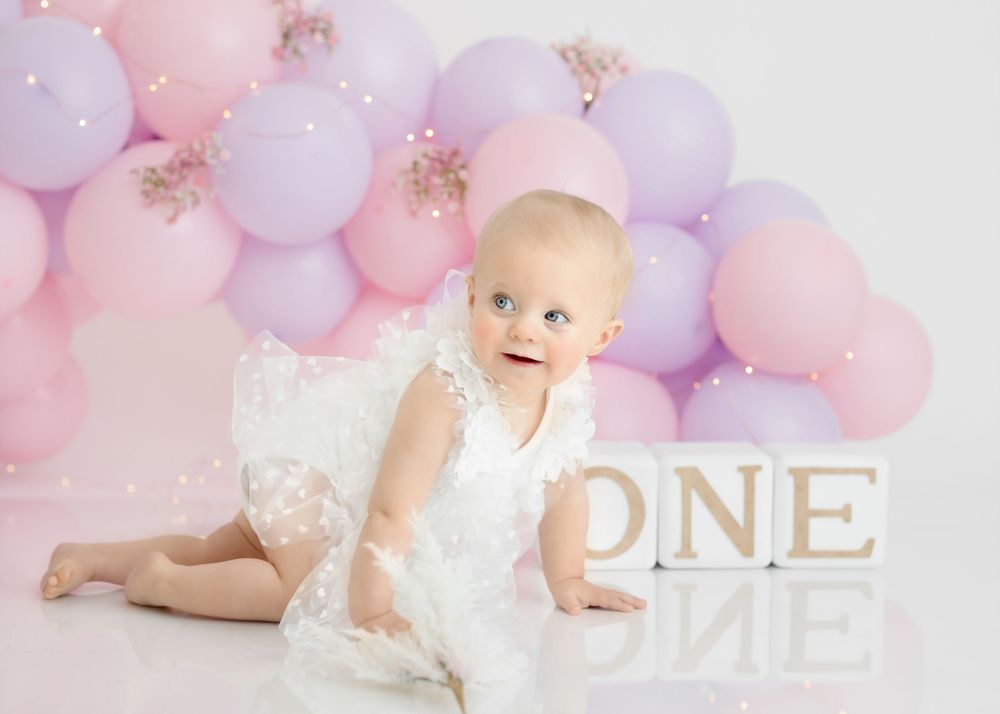 First birthday portrait with white lace outfit and pink and purple balloon backdrop.