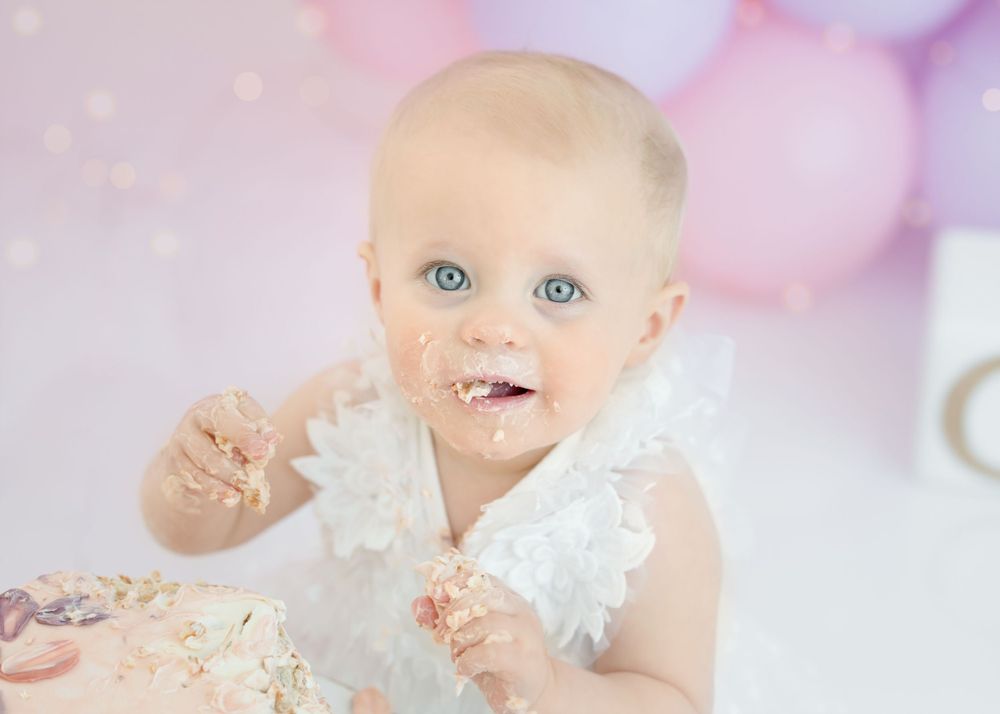 Close-up cake smash portrait with pink frosted cake and pastel balloon backdrop.