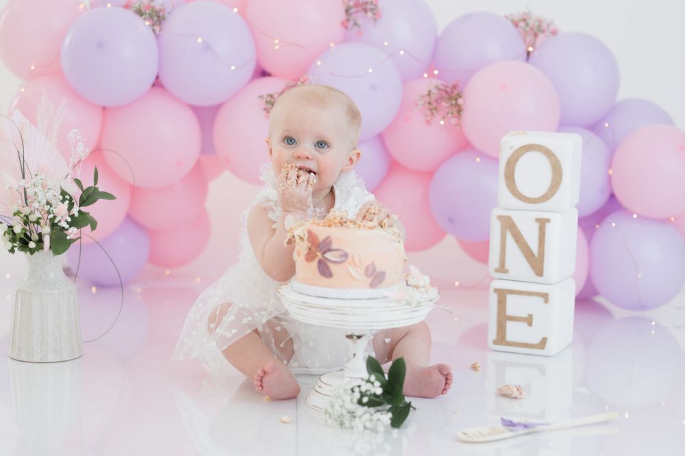 First birthday cake smash scene with pink and purple balloons, white cake stand, and gold letter blocks spelling ONE.