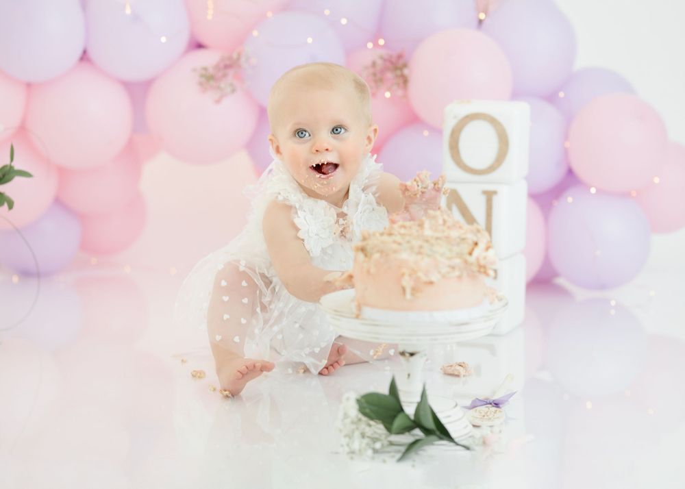 First birthday photo setup with pink and purple balloon arch and lace dress against white background.