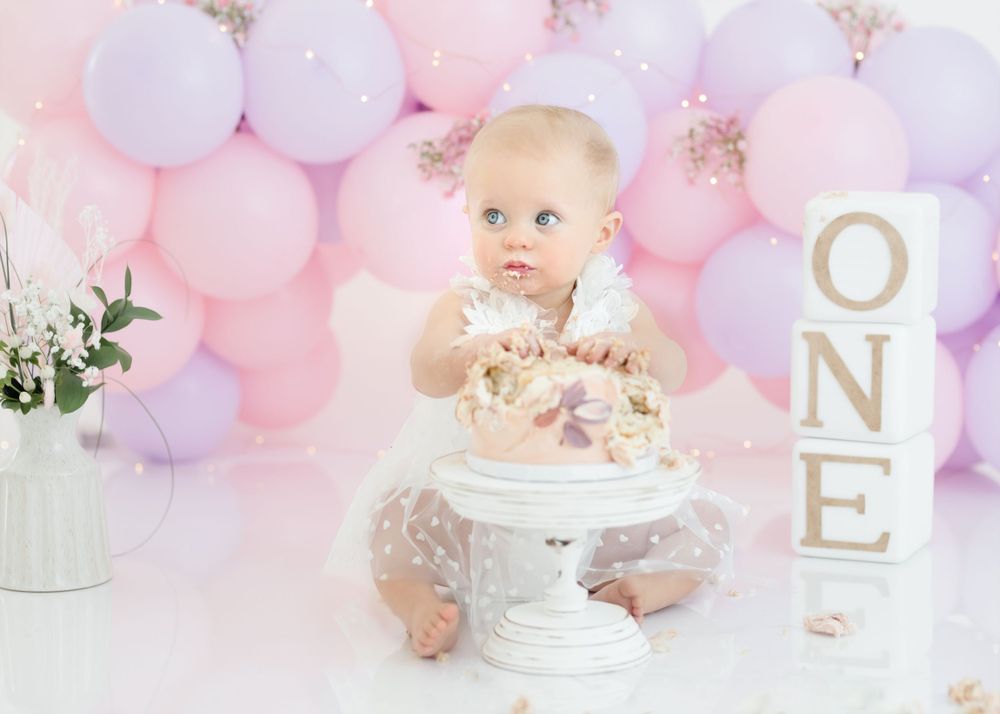 Baby enjoying birthday cake on white pedestal against pink and purple balloon backdrop.