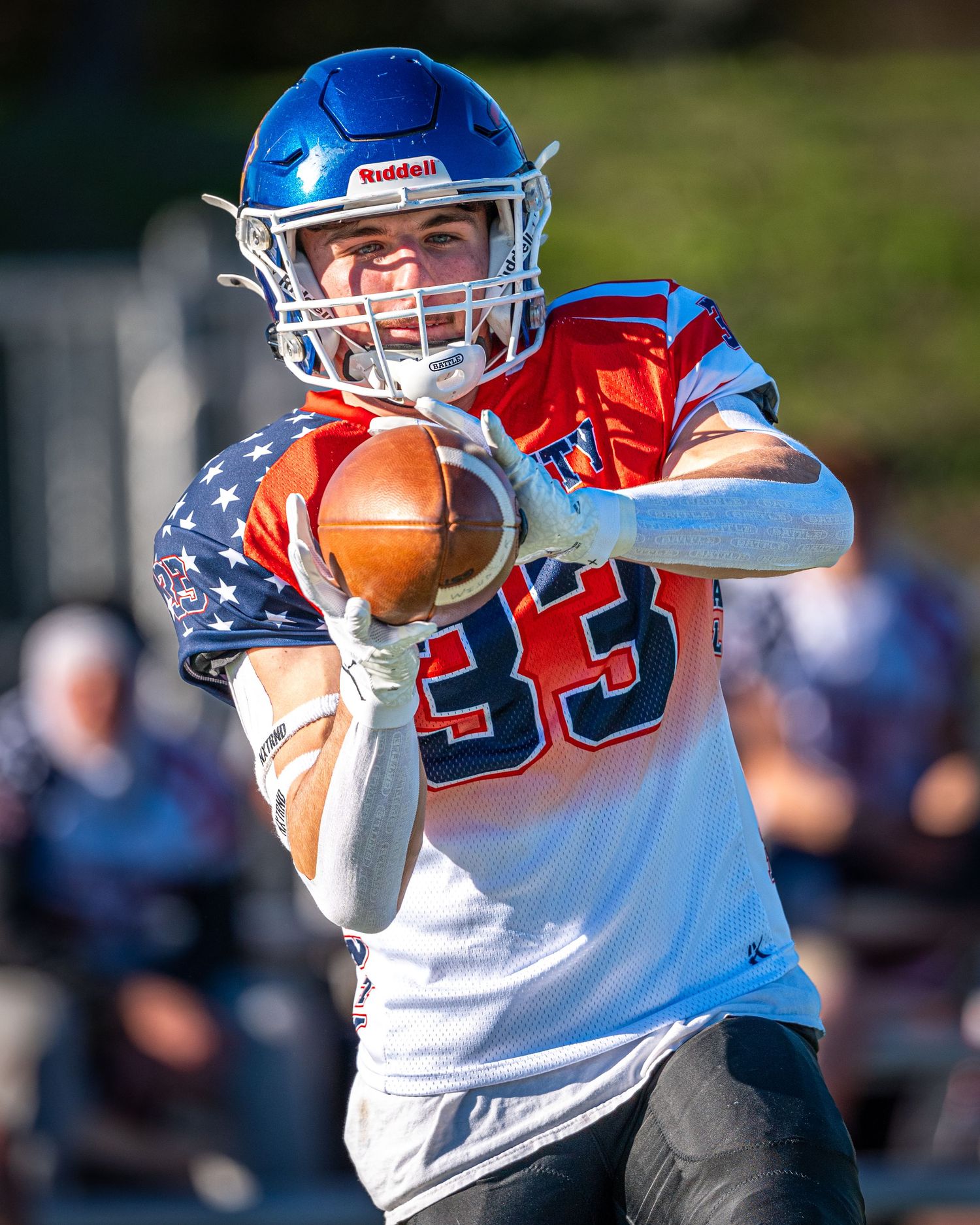 Youth football player in blue helmet reaches to catch an American football in red and white uniform during game action.