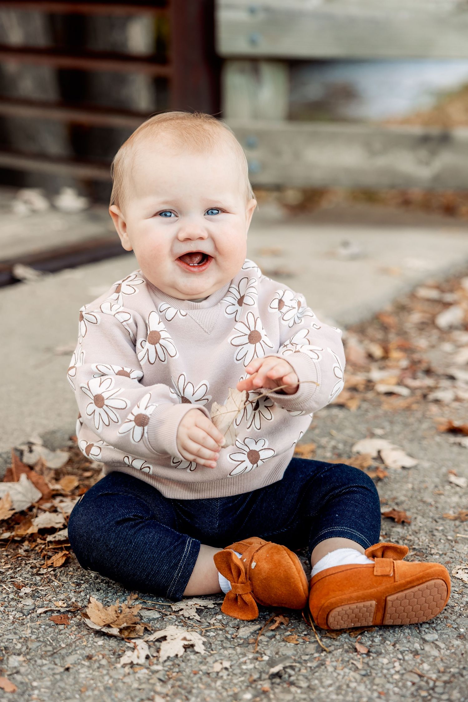 A baby in a floral sweater and denim sits on the ground smiling while wearing brown leather shoes. Michigan Detroit Novi Photographer