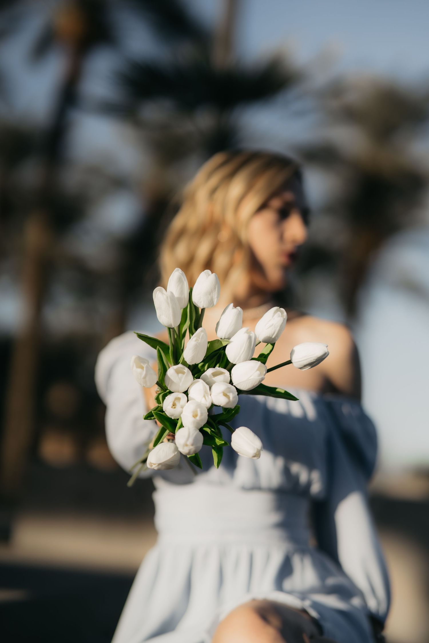Woman in white dress holds fresh bouquet of white tulips in outdoor setting with blurred background.
