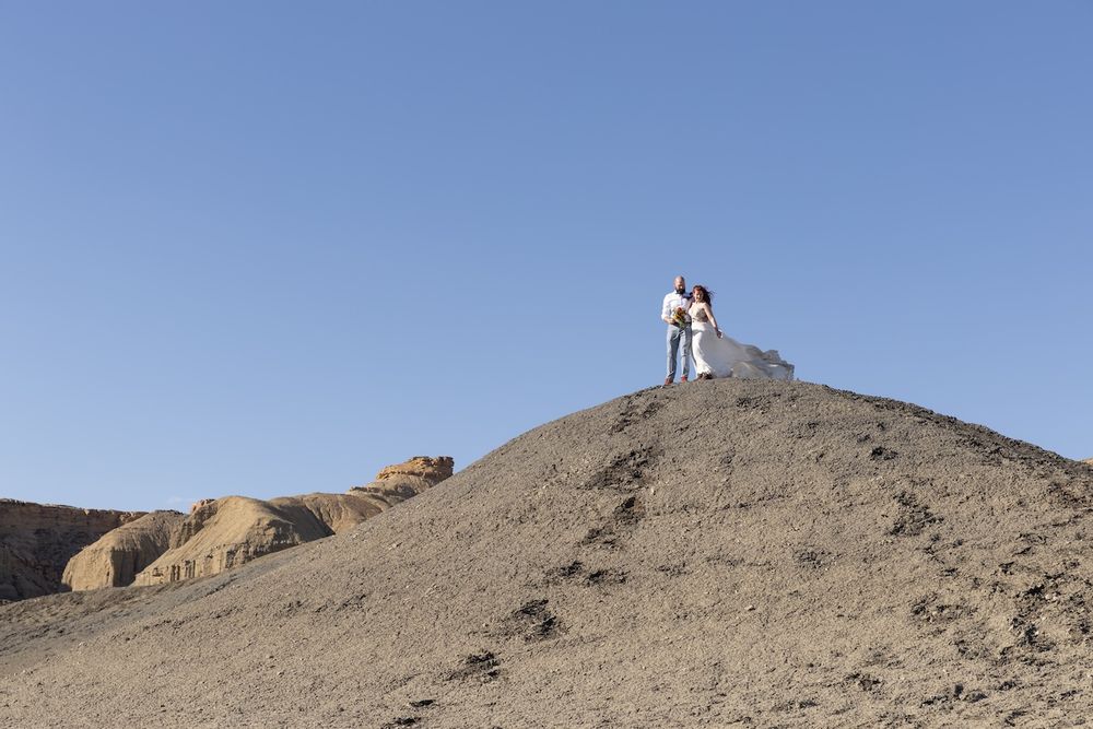 A couple in wedding attire stands atop a desert mountain ridge against a blue sky backdrop for their photoshoot.