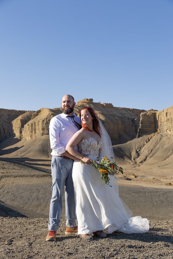 A couple in wedding attire poses together against dramatic desert cliffs and rugged terrain at sunset.