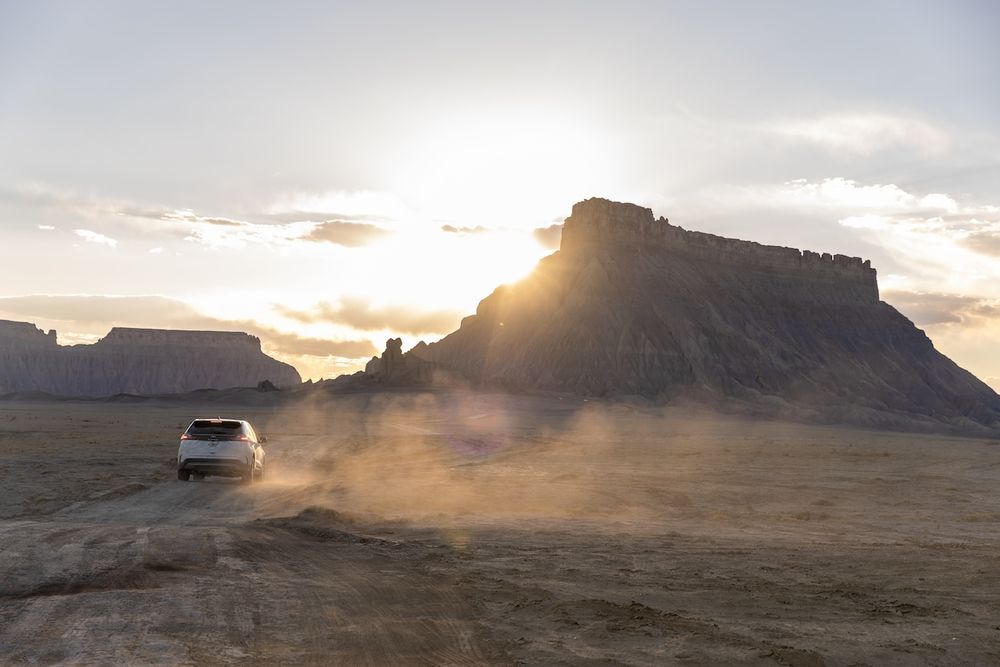 SUV kicks up dust on a desert road at sunset with dramatic mesa formations silhouetted against the golden sky.
