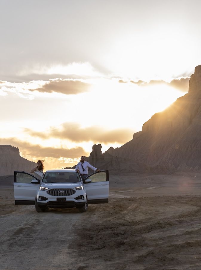 White car with doors open parked in desert landscape during dramatic sunset with rocky cliffs in background.