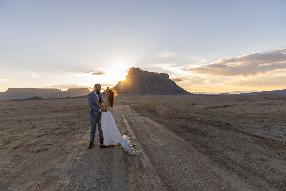 Silhouettes stand against a dramatic desert sunset with mountains and rock formations in the background.