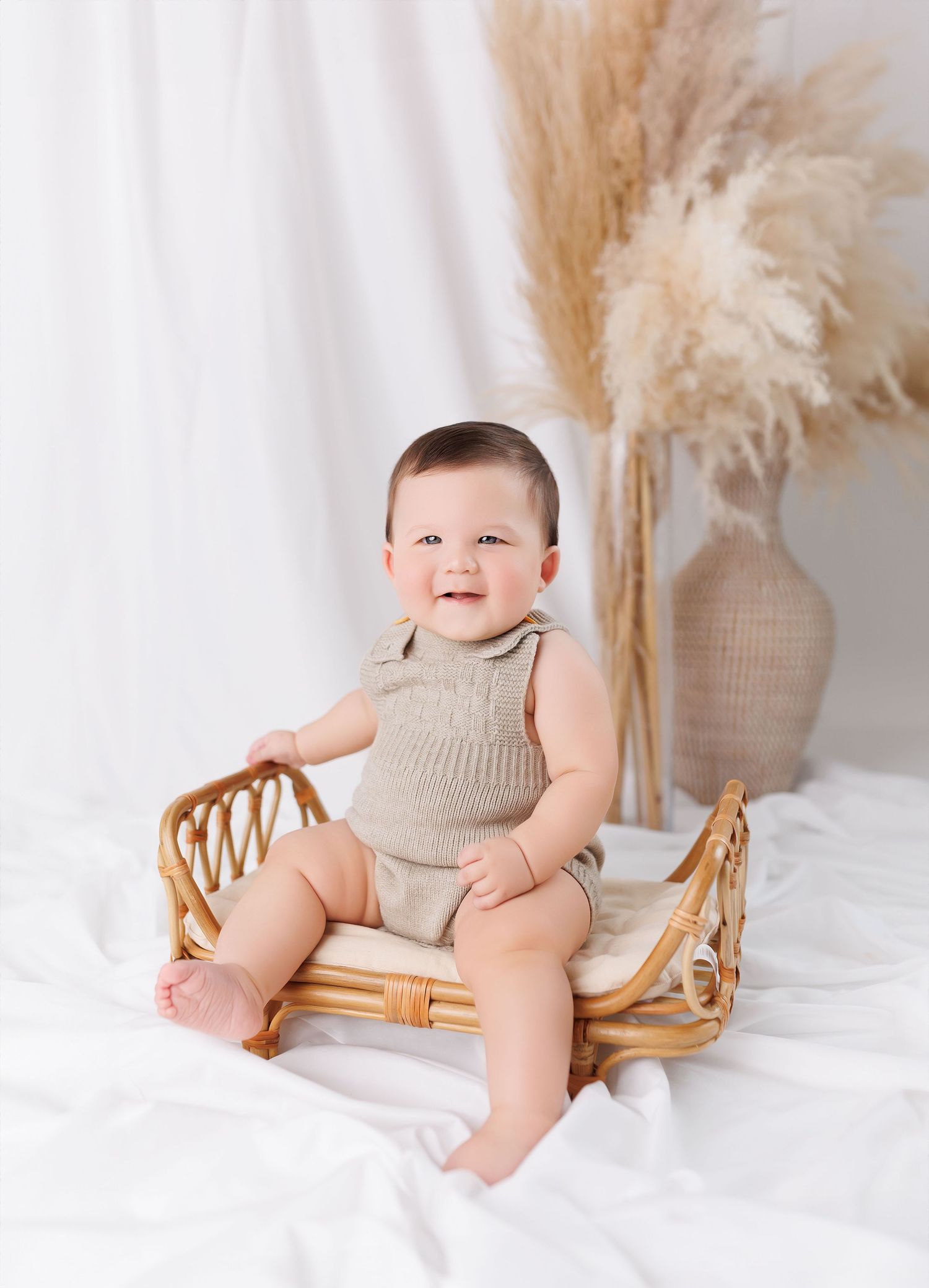 Baby in knit romper poses on rattan chair with neutral boho decor and pampas grass background.