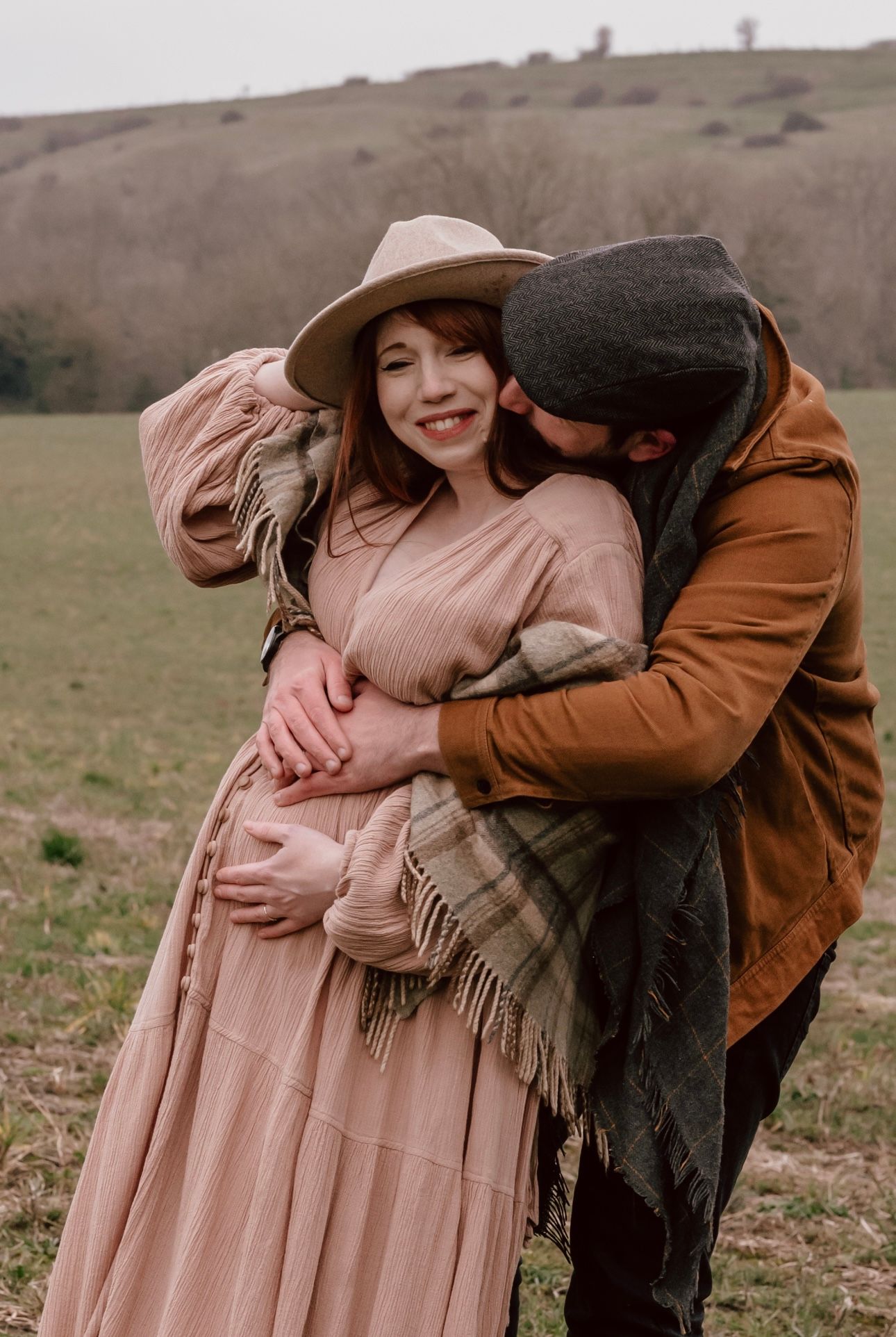A couple embrace in the wiltshire countryside. He hugs her from behind, cradling her pregnant stomach with tenderness.