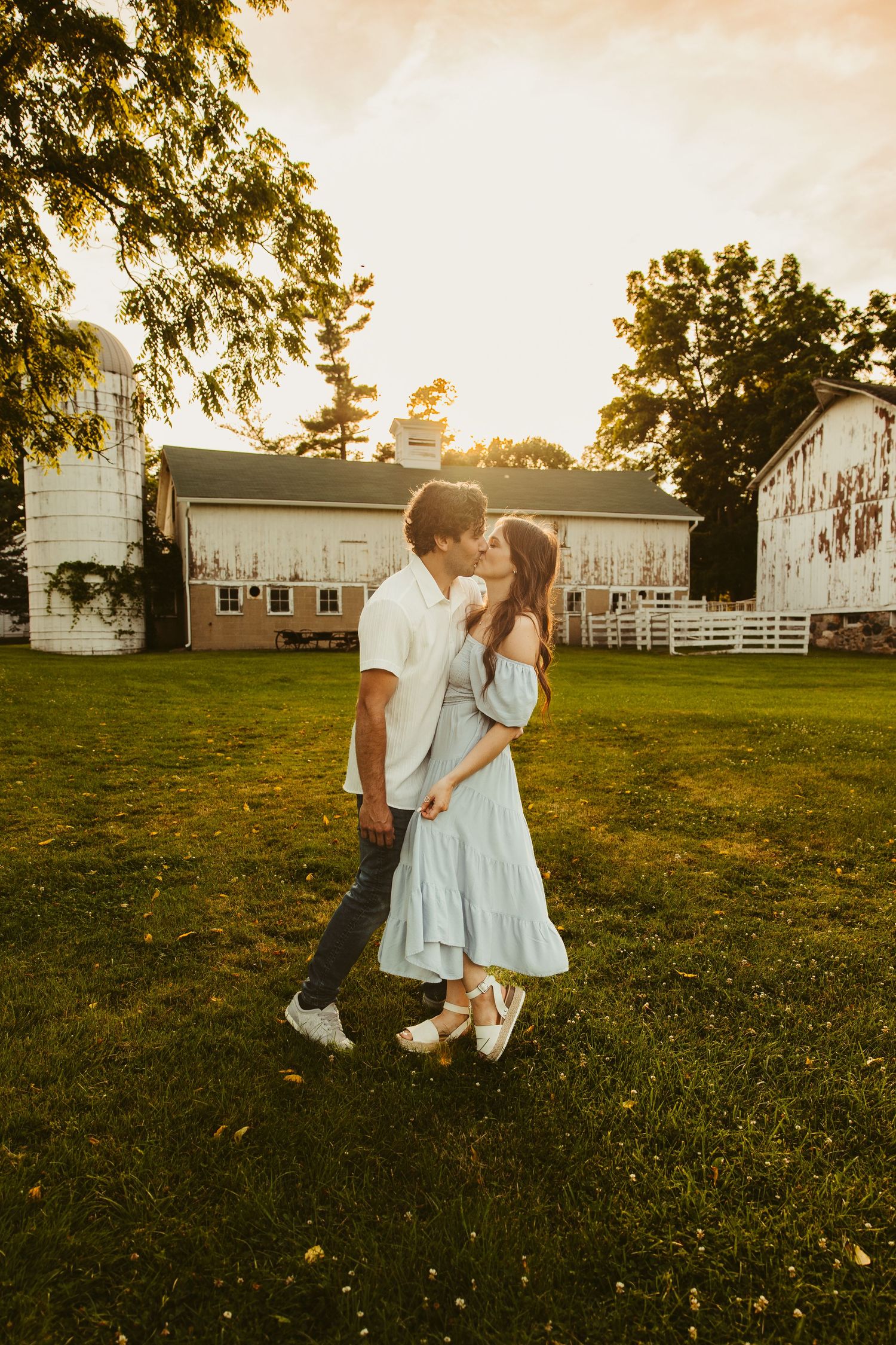 A couple shares a romantic moment at sunset near a rustic barn with white fences and golden light filtering through trees.
