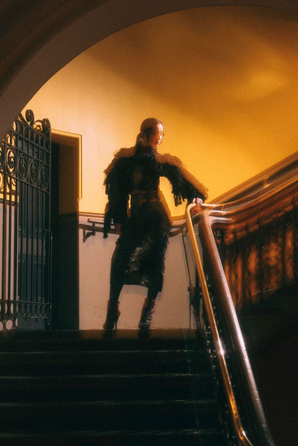 Silhouette on staircase wearing feathered black ensemble in atmospheric lighting.