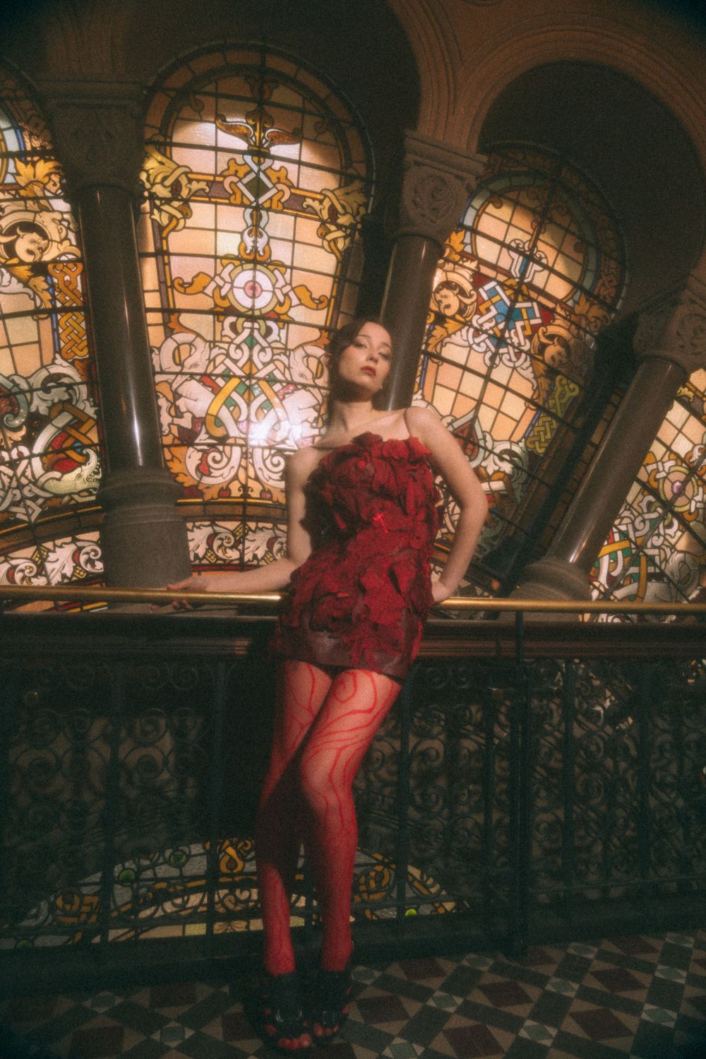 Figure in red dress posing against ornate stained glass windows in vintage setting.