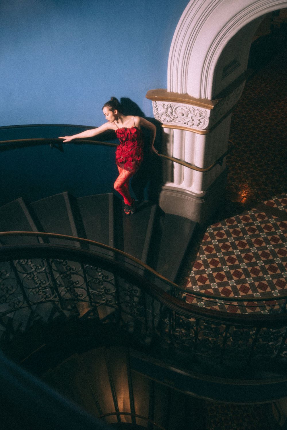 A dramatic shot of a figure in a red dress posing on an ornate staircase with classical architecture.