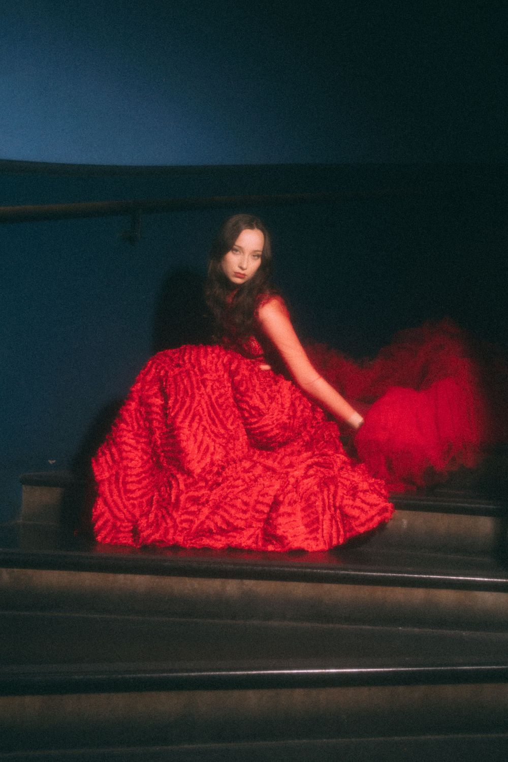 Figure in a flowing red dress posed on dark stairs with artistic lighting.
