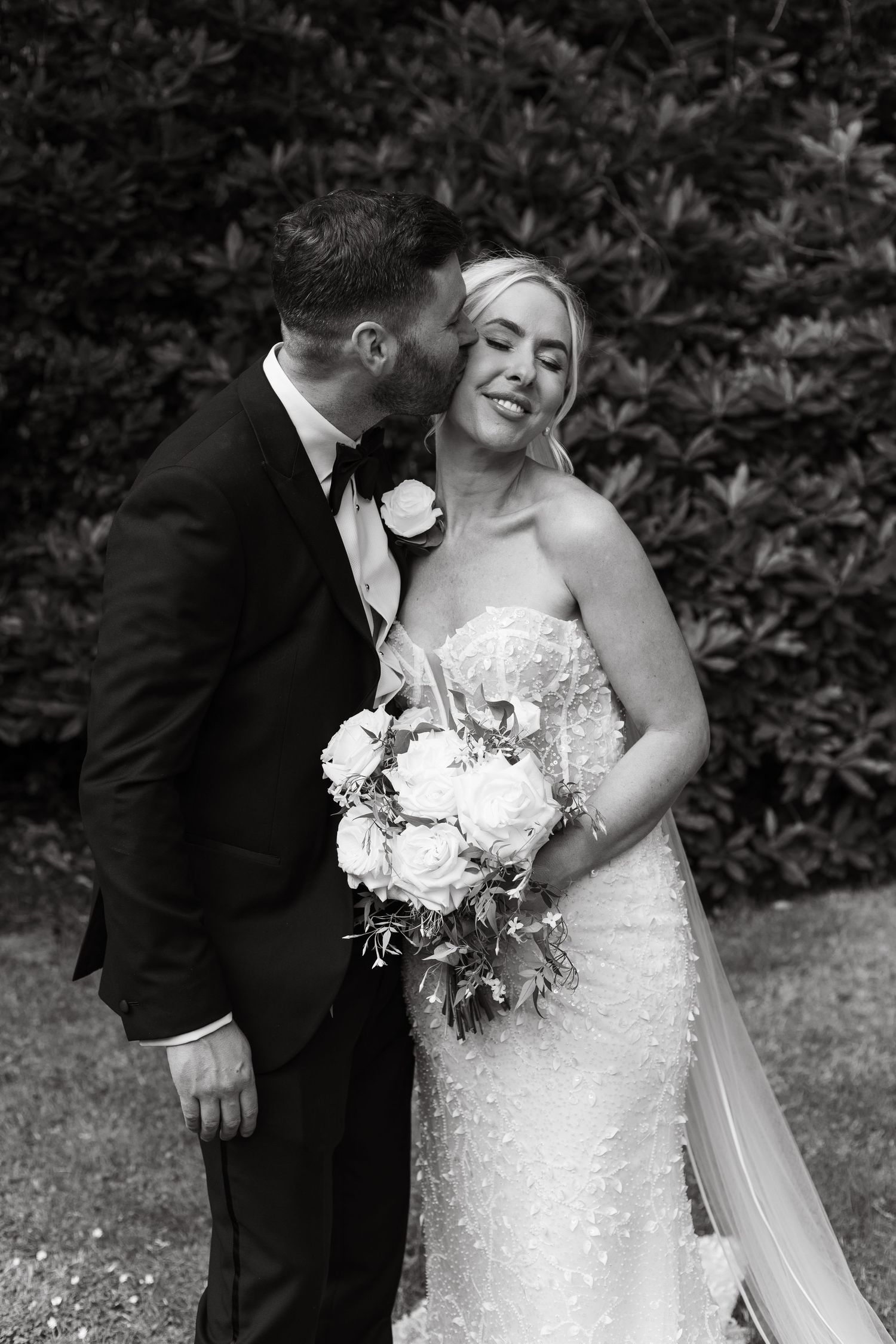 Romantic black and white wedding portrait of a couple with white floral bouquet in an outdoor garden setting.