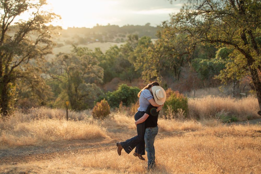 Couple embracing in golden sunlit field with oak trees in background.