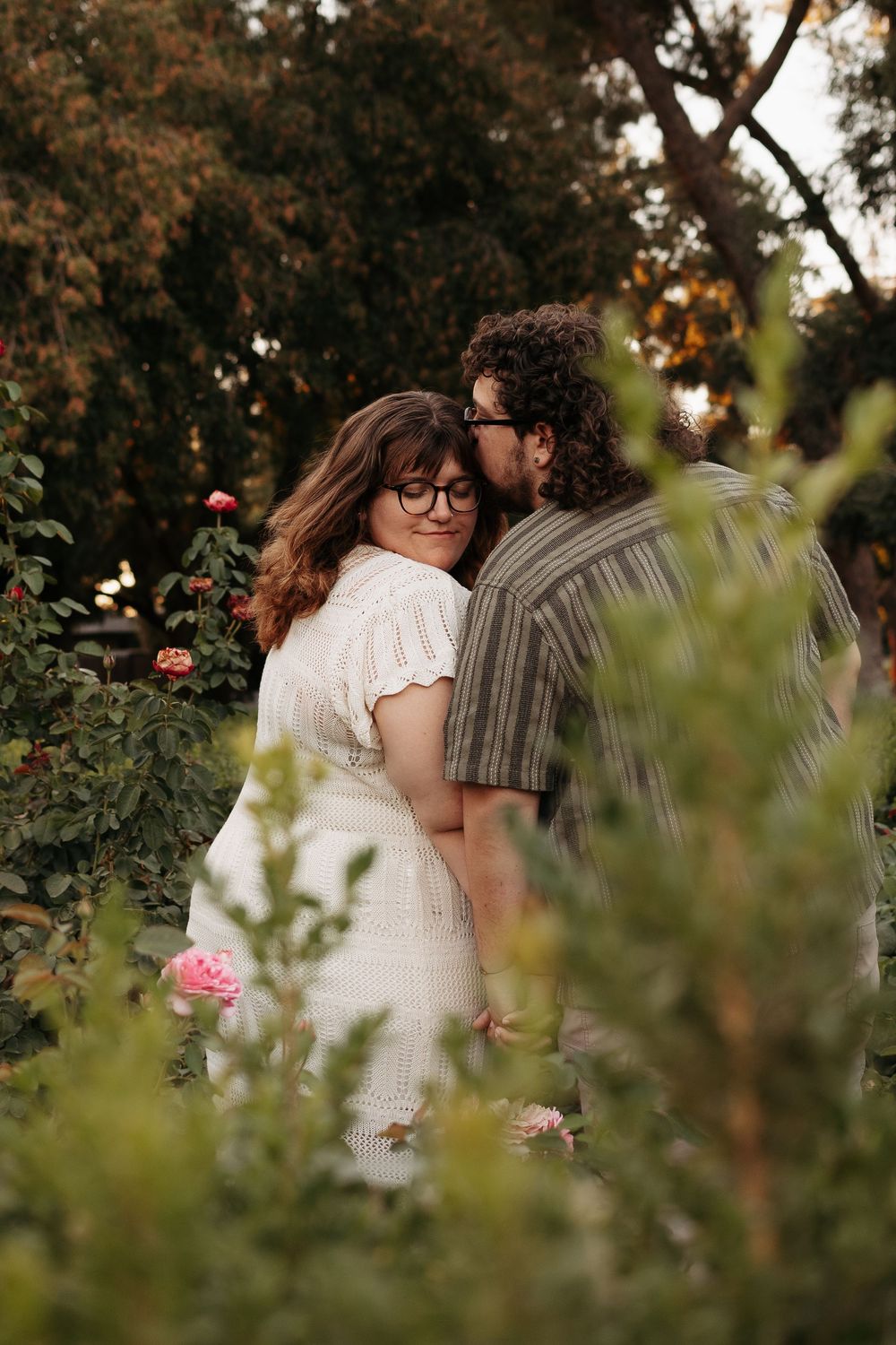 A romantic couple shares intimate moments among lush greenery and trees during a sunset photoshoot.