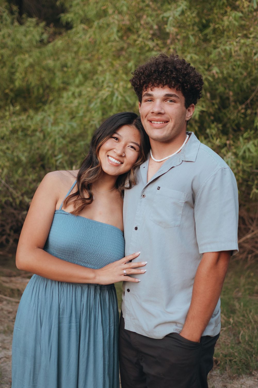 A couple poses together outdoors, with one wearing a blue dress and the other in a light colored polo shirt.