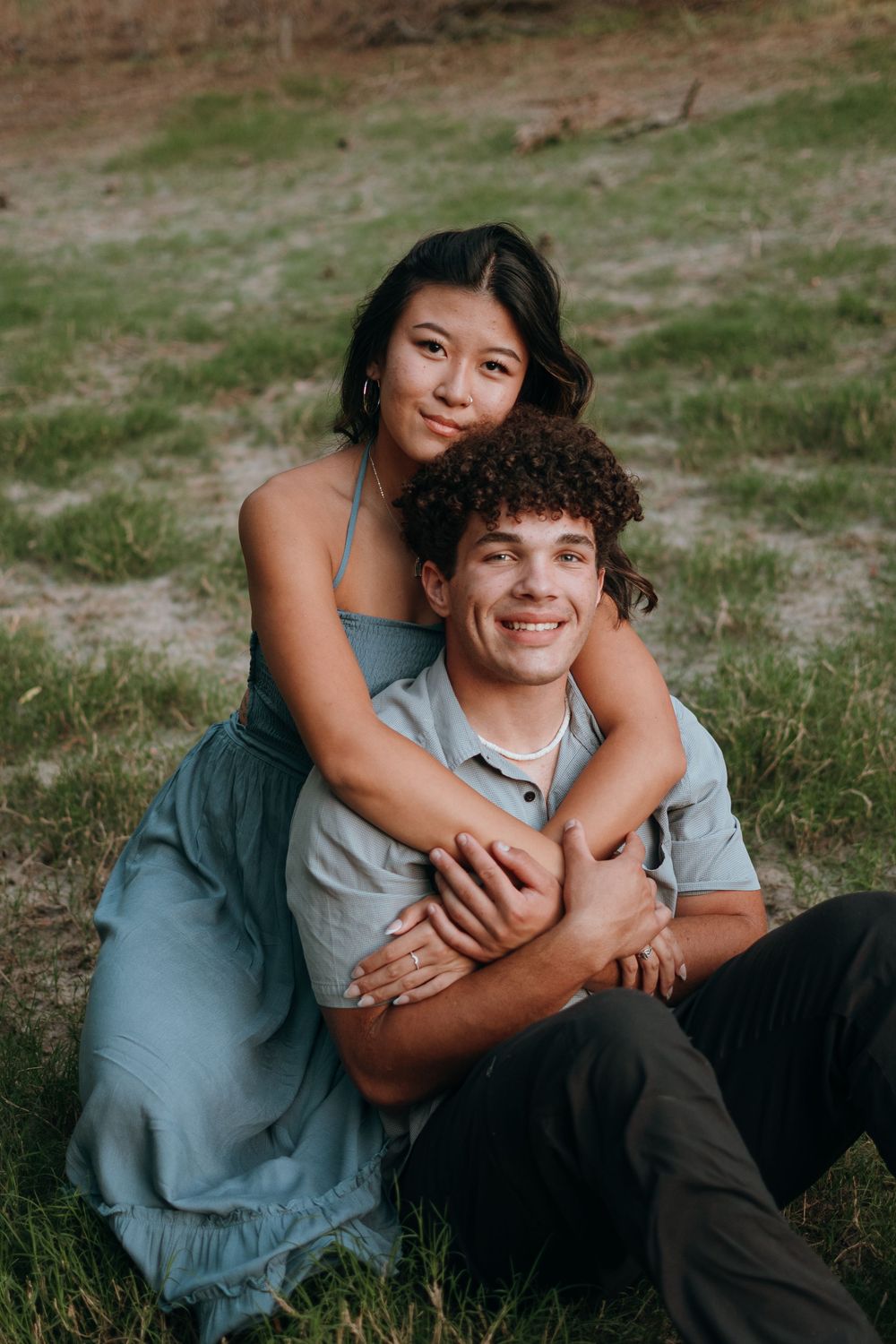 A couple sits together on grass, sharing an intimate embrace while wearing casual clothing in soft evening light.