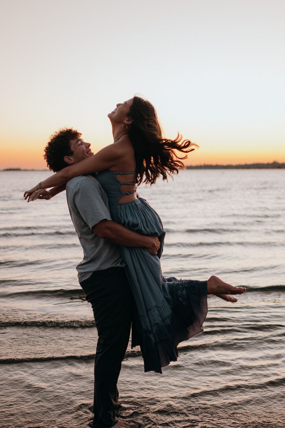 A couple dances playfully in the ocean waves during sunset, creating a romantic silhouette against the golden sky.