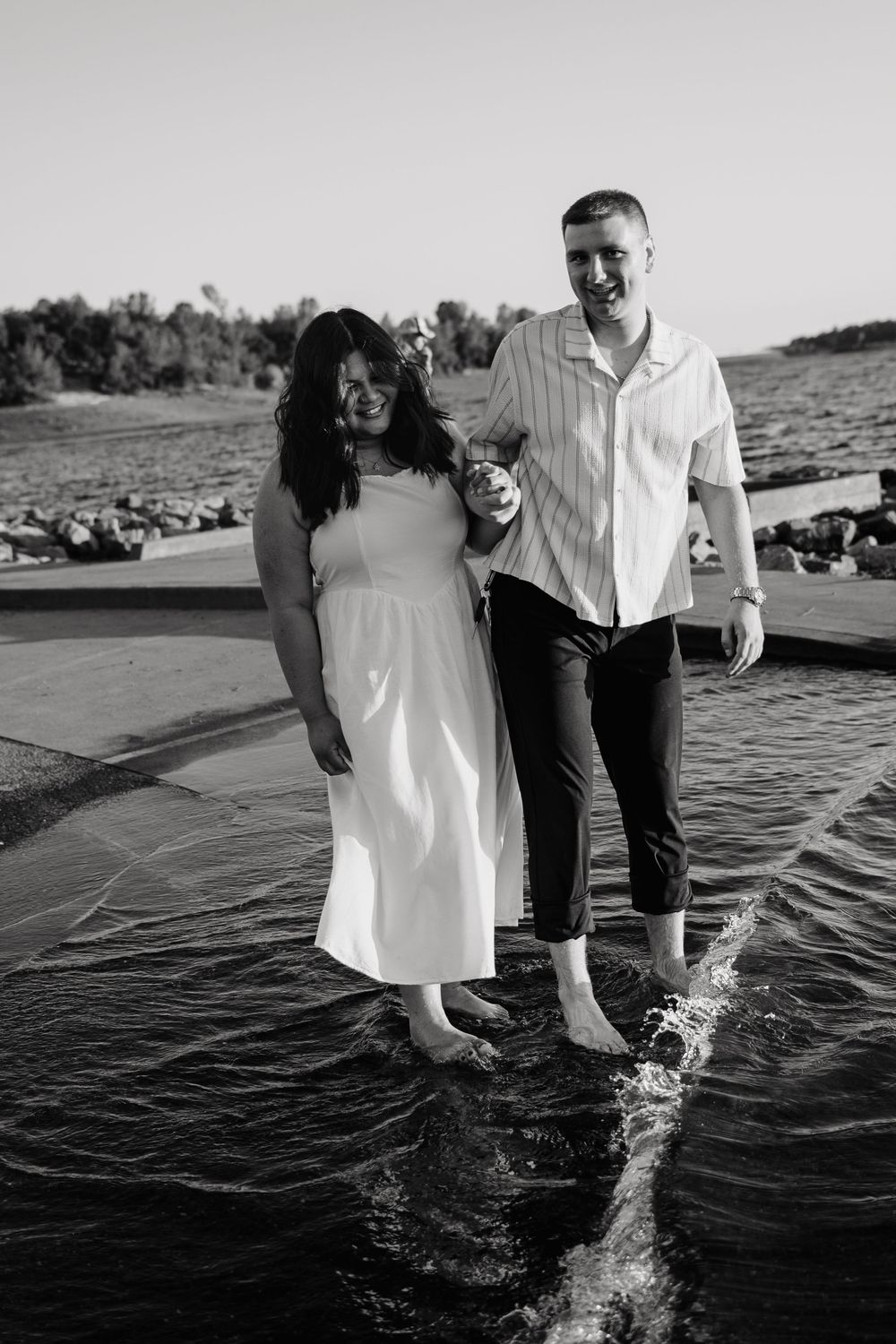 Black and white photo of couple splashing and playing in shallow water at beach.
