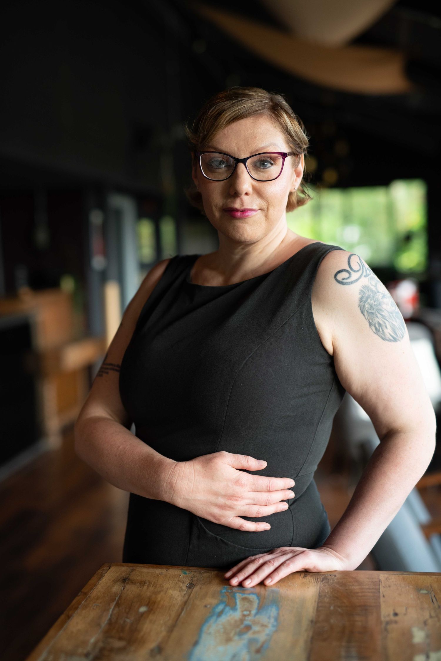 Portrait of woman in black sleeveless top with visible arm tattoo leaning on wooden table.