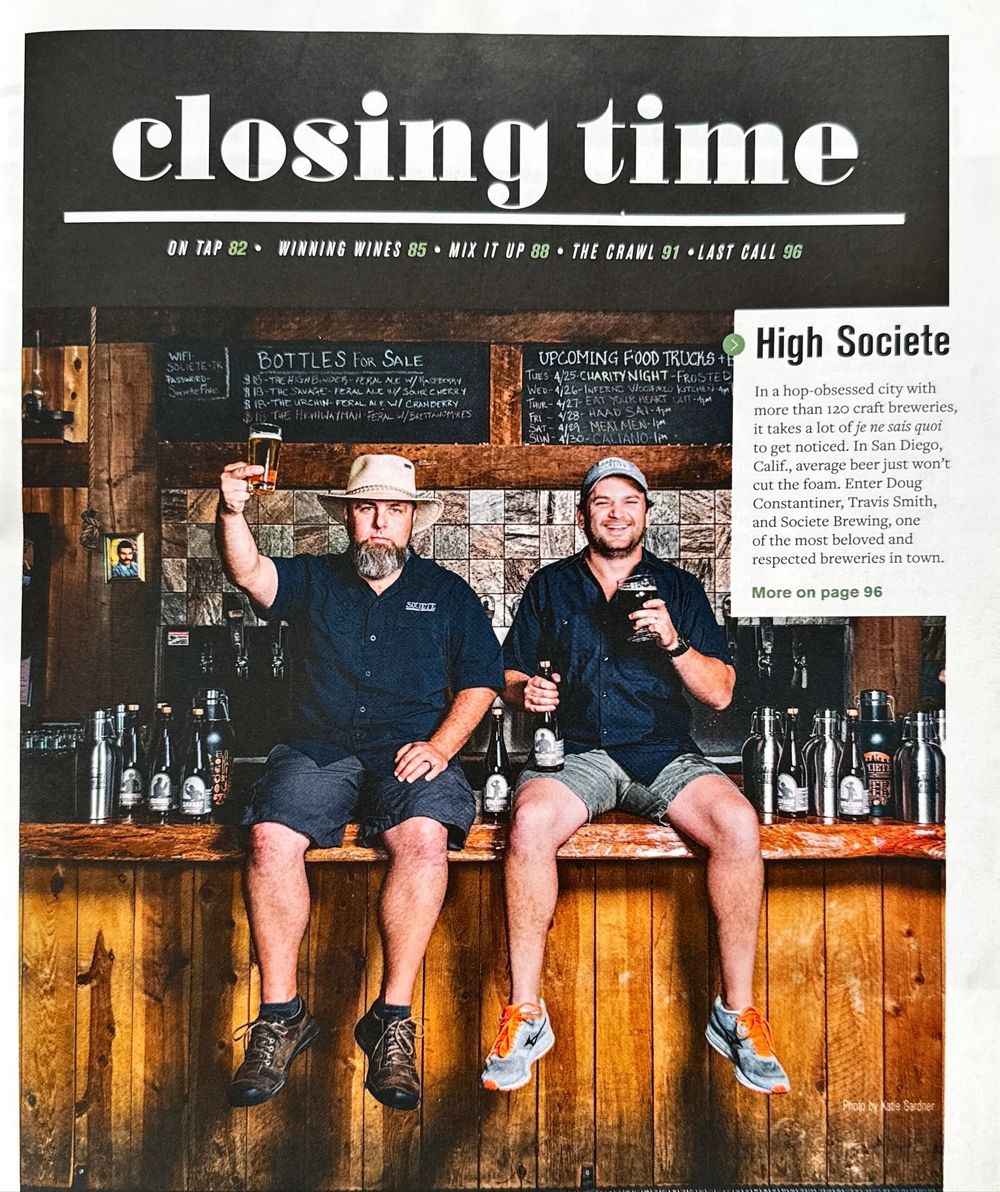 Four people sit on a wooden bar counter with their legs dangling while holding drinks in a rustic pub setting.