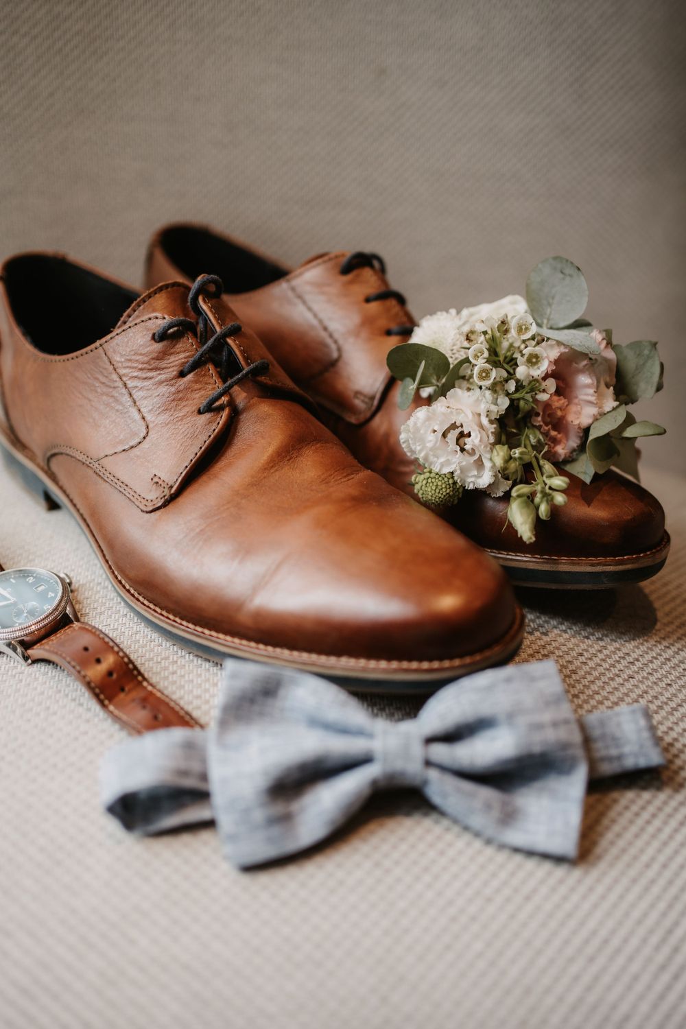 Brown leather dress shoes with gray bowtie and small floral boutonniere on beige fabric.