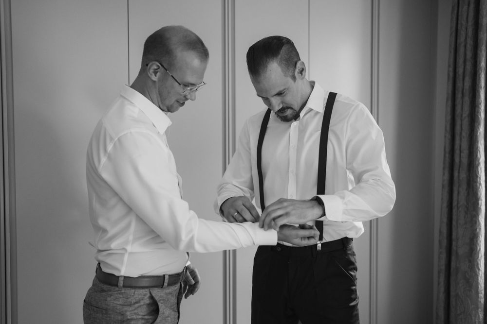 Two men in white shirts and suspenders prepare during pre-wedding moments.