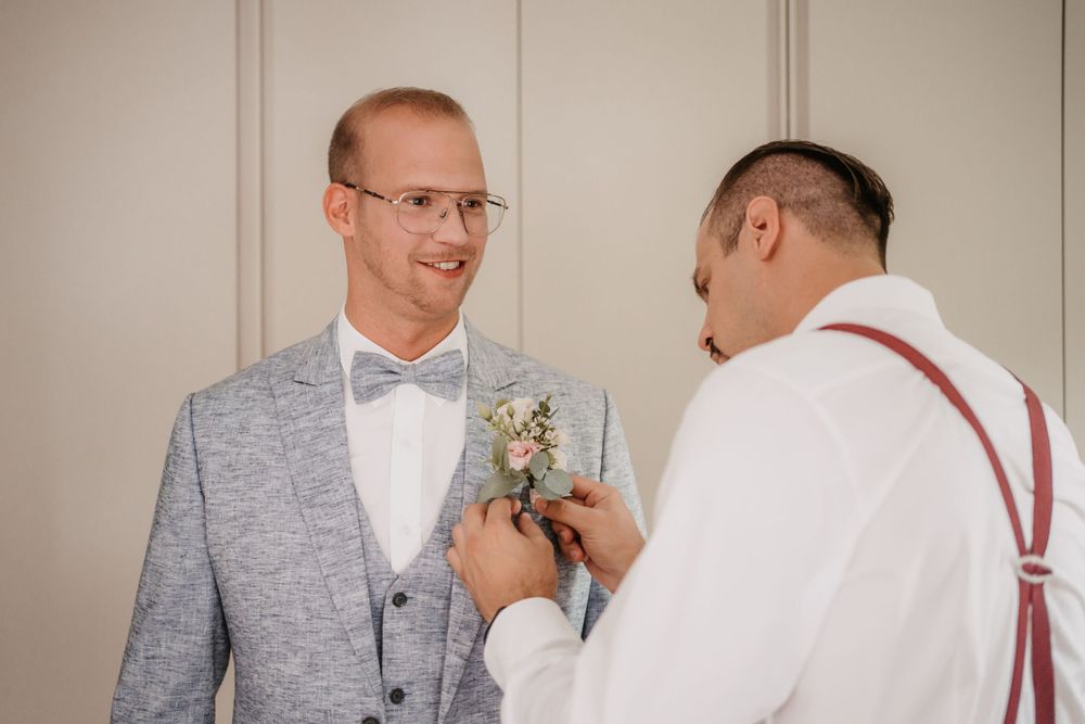 Groomsman helps adjust boutonniere on groom's grey suit lapel.