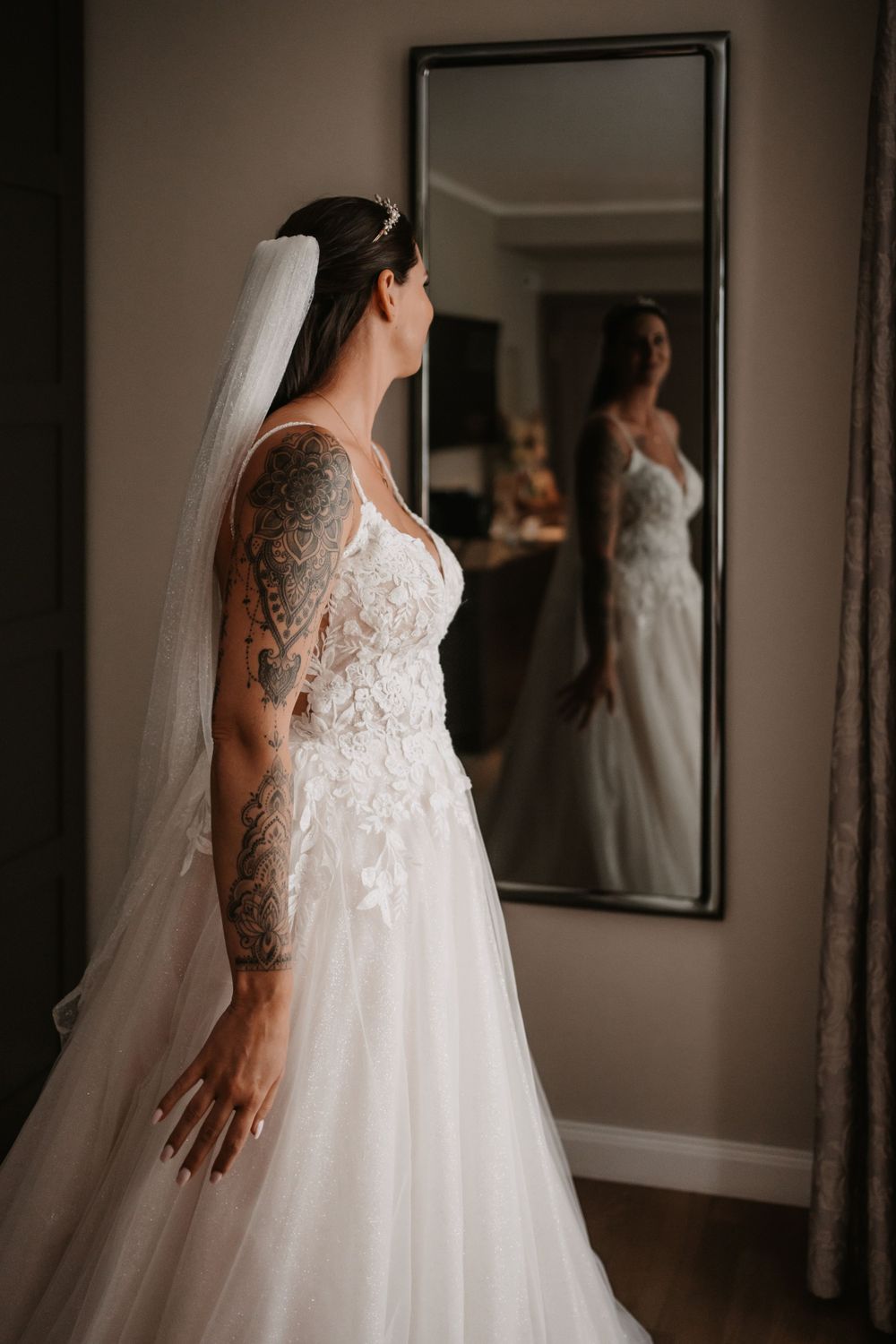Elegant bride in lace gown with tattoo admiring reflection in full-length mirror.