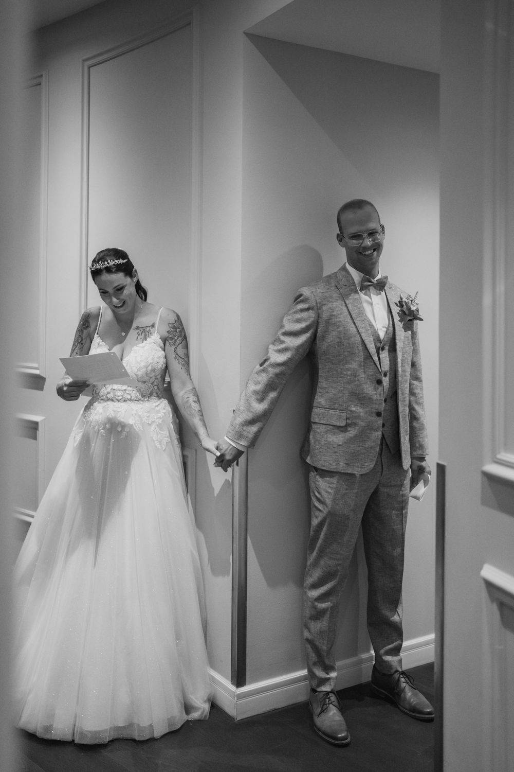 Black and white photo of couple doing first look while holding hands around corner.