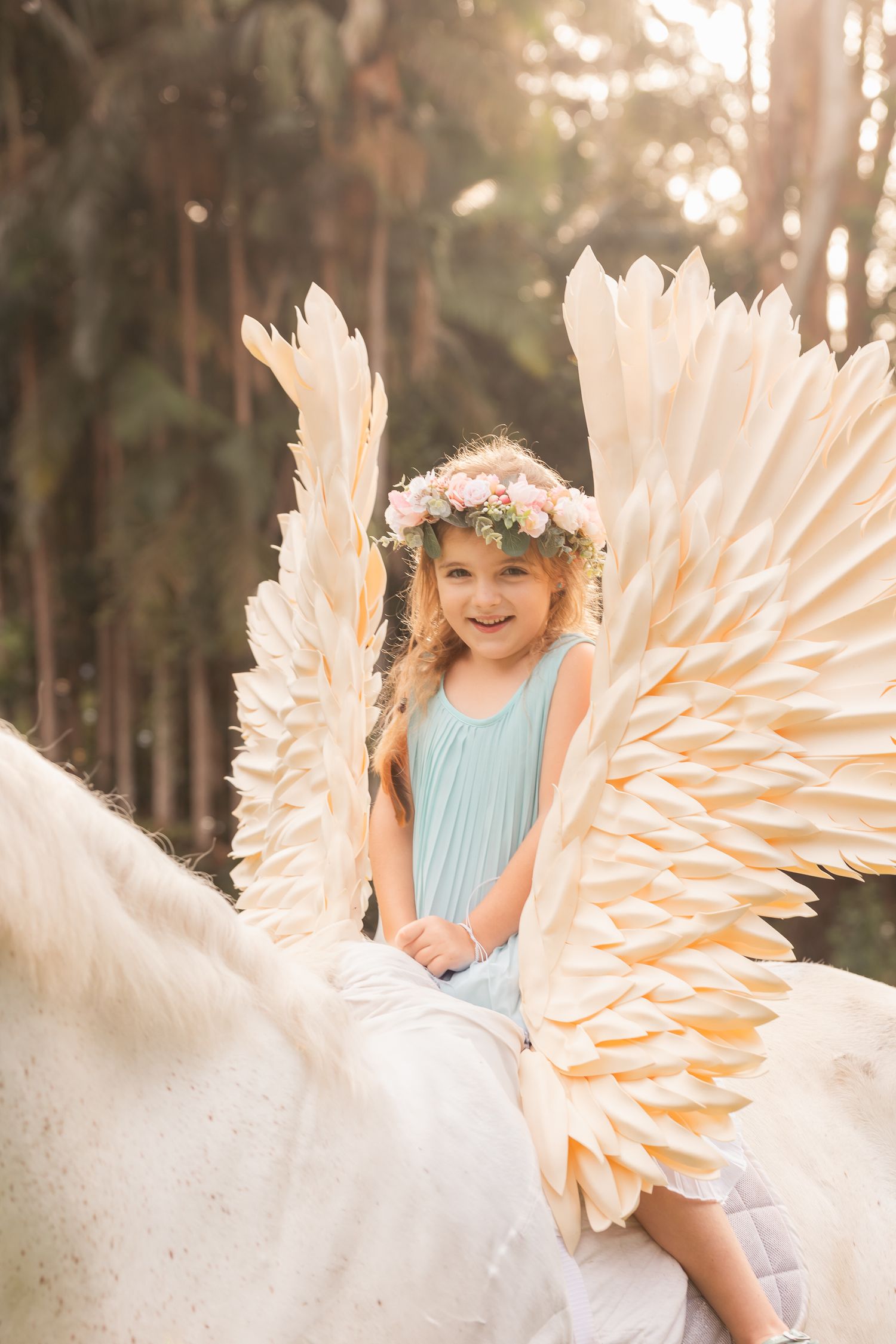 Whimsical outdoor portrait with large cream-colored angel wings and floral crown.