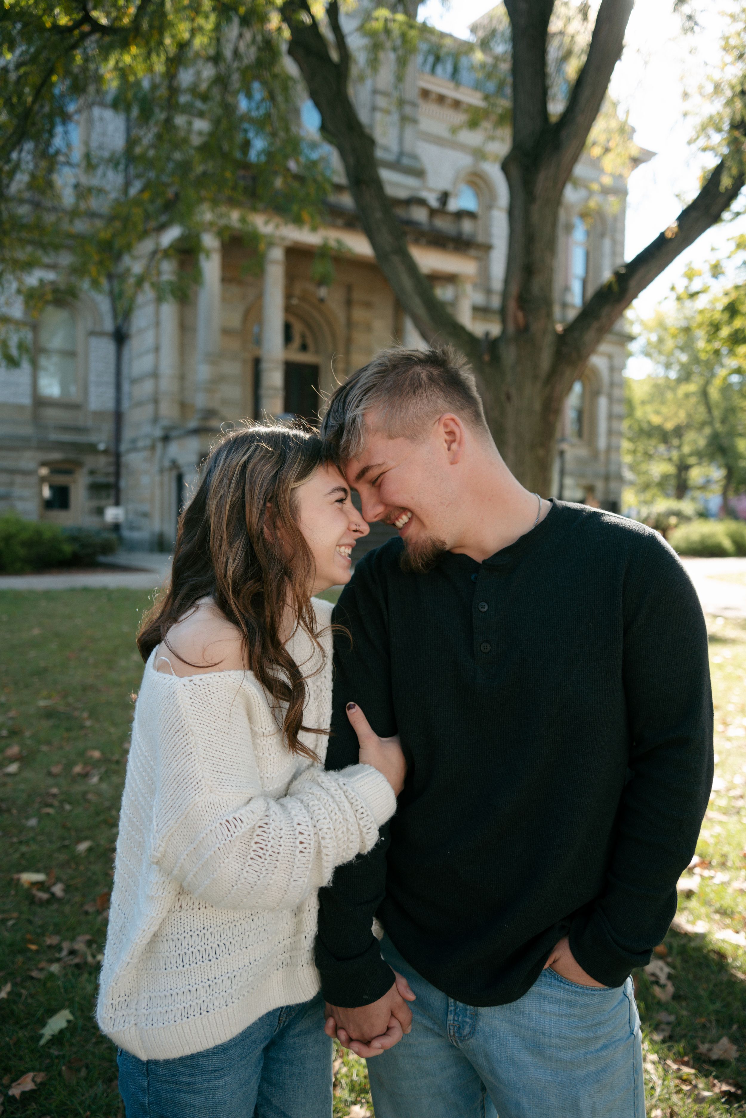 Smiling couple holds hands and touches foreheads near a large tree outside a historic building.