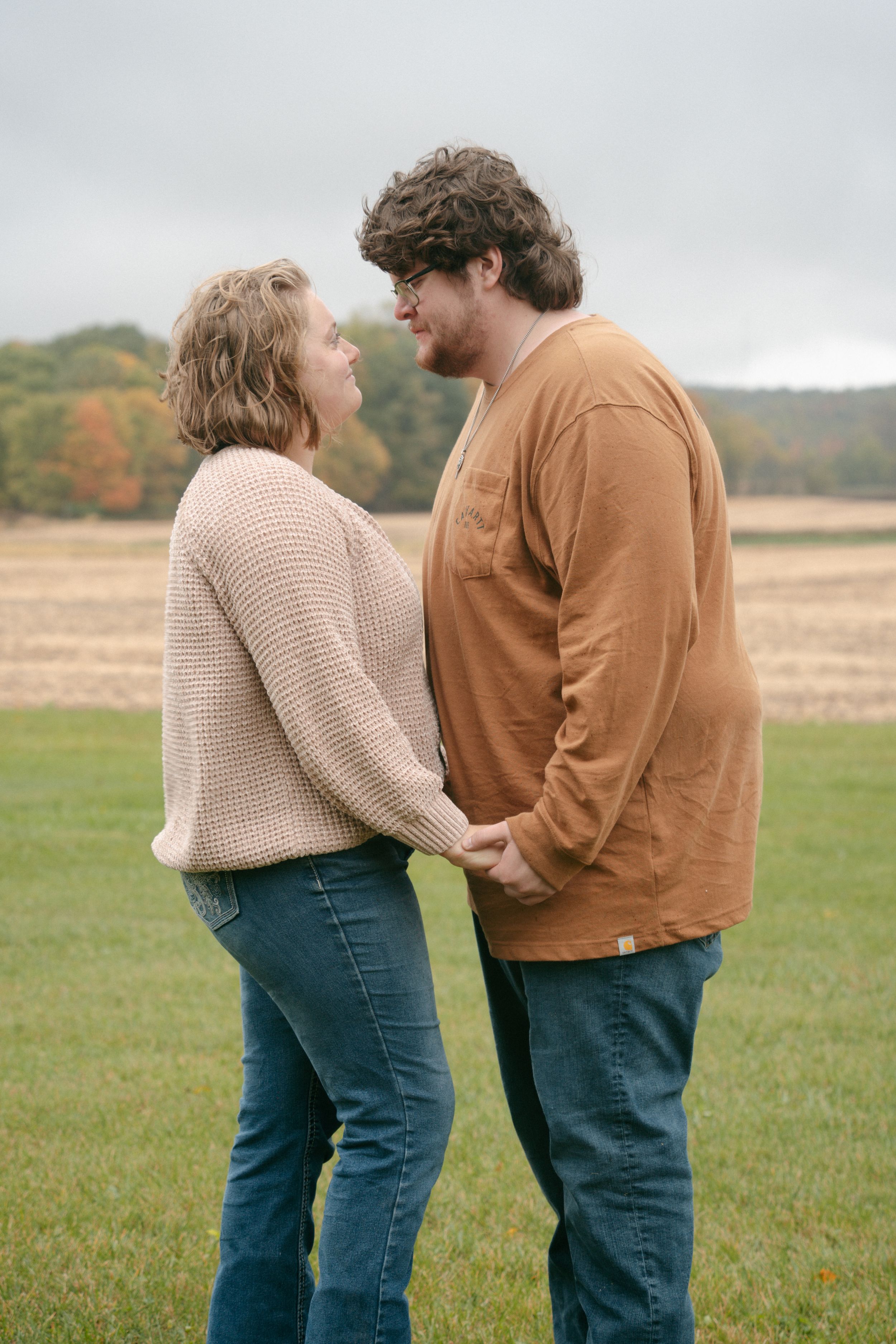 Couple holding hands face each other in an open field with autumn foliage in the background.