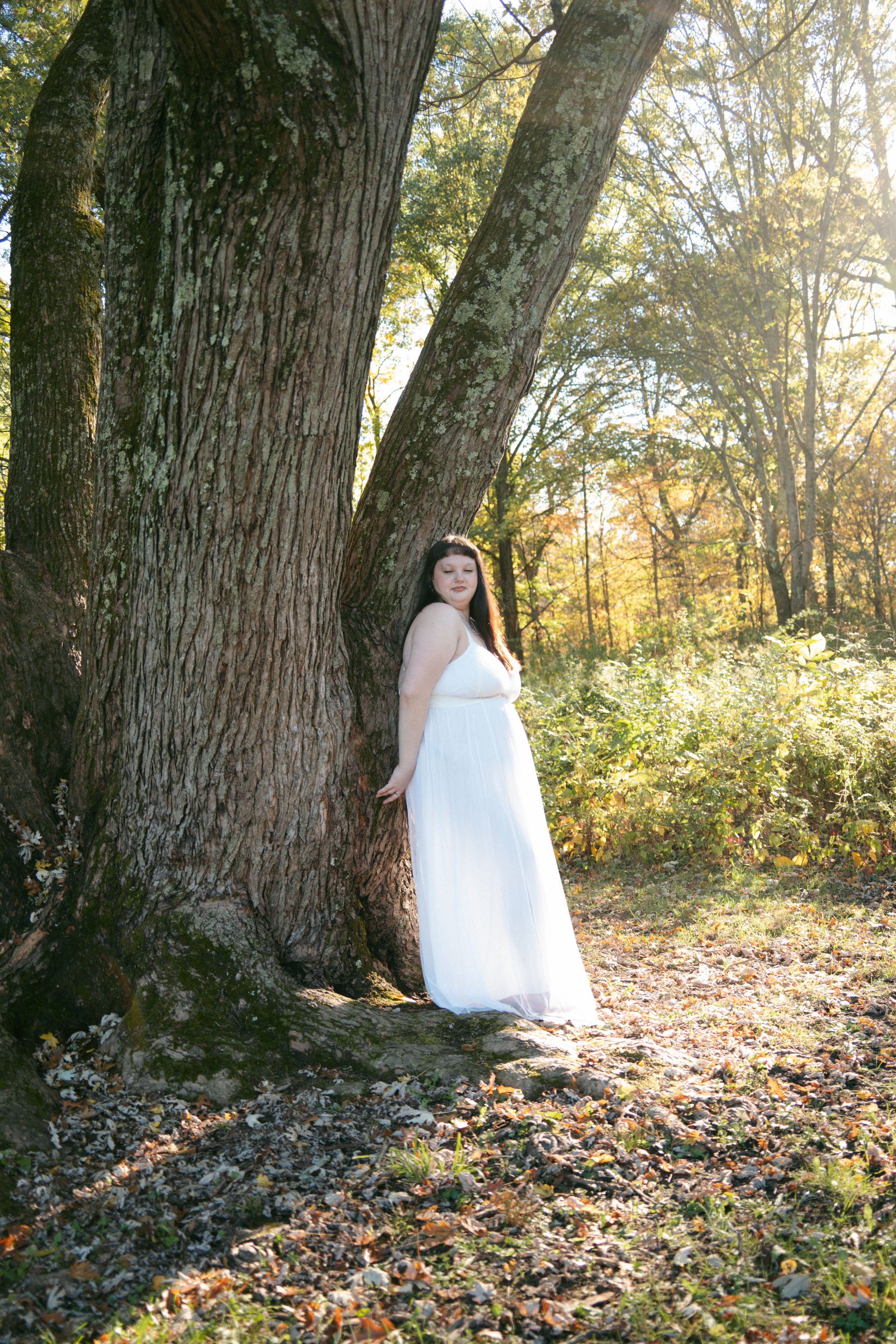 Woman in white gown leans against a large tree trunk in a sunlit autumn forest.