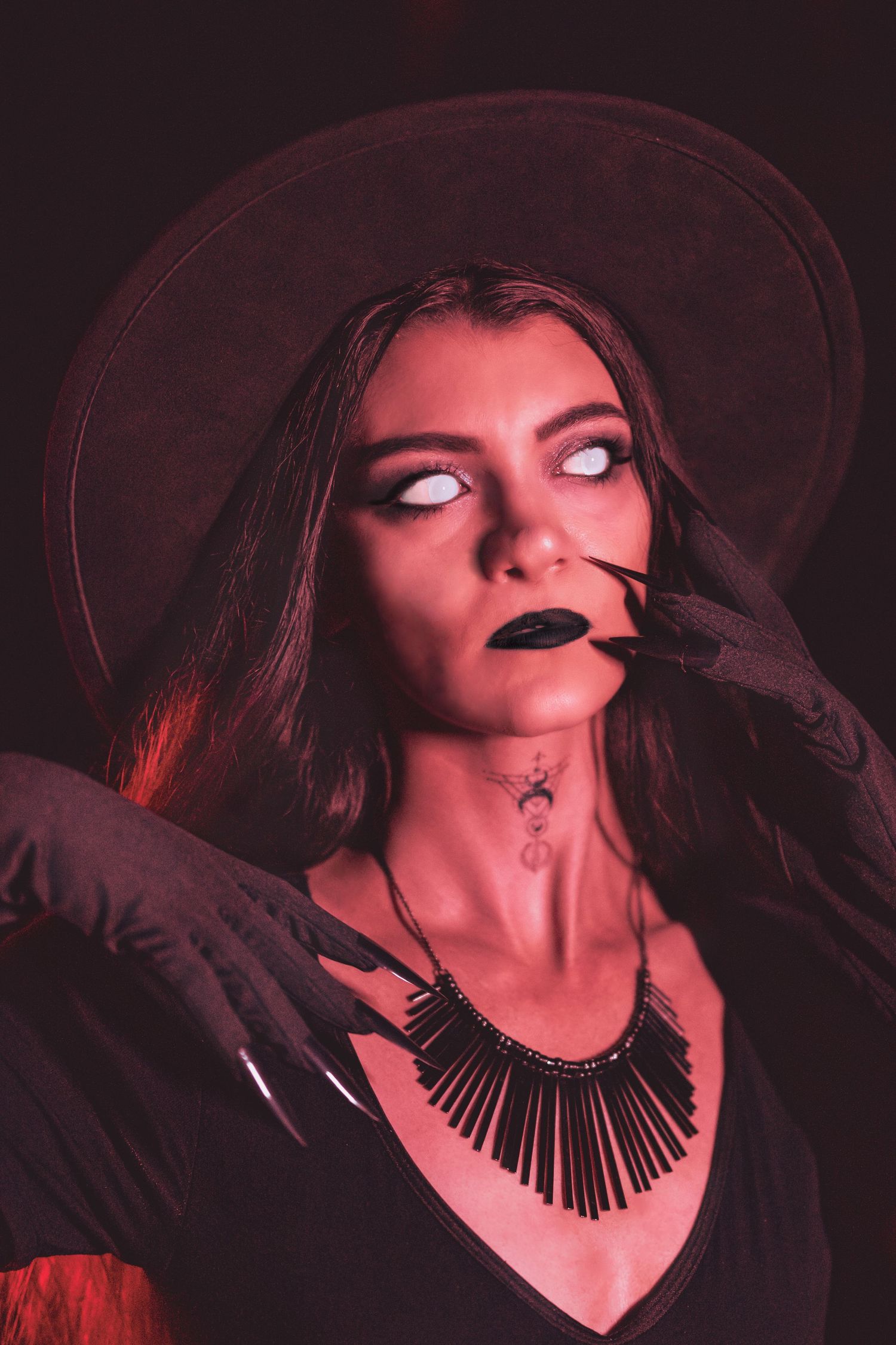 A woman in dramatic witch Halloween costume with black hat, necklace, and white contact lenses.