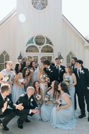 Wedding party in light blue dresses and black tuxedos laughs together in front of white chapel building.