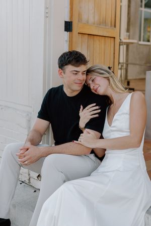 A couple sits close together on white steps, sharing an intimate moment in casual and formal attire.